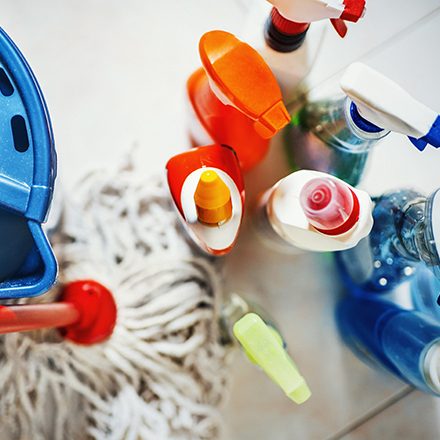 Closeup top view of unrecognizable home cleaning products with blue bucket and a mop on the side. All products placed on white tiled bathroom floor.